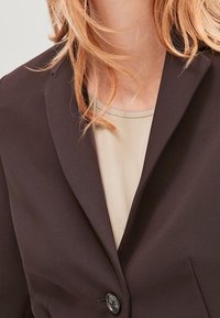 Kobieta nosząca ciemno-brązową marynarkę na beżowej bluzce, z luźnymi blond włosami częściowo widocznymi wokół szyi i ramion.