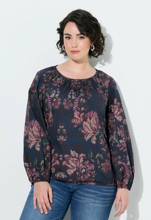 Mujer con cabello corto, rizado y oscuro, que lleva pendientes de aro plateados y una blusa oscura de manga larga con estampado floral combinada con vaqueros azules sobre un fondo blanco.