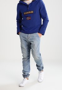 Blå letvægts hættetrøje med en frontlomme med lynlås, med et logo og flagdetalje. Kombineret med lyseblå jeans og hvide sneakers.