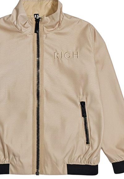 Giacca beige con cerniera e collo alla coreana, dettagli con cerniera nera, polsini e orlo a coste, e la scritta "RICH" ricamata sul petto.