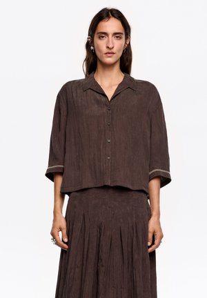 Camisa de tela marrón arrugada con cuello, mangas cortas y cierre de botones. Detalle notable de costura en el dobladillo de la manga.