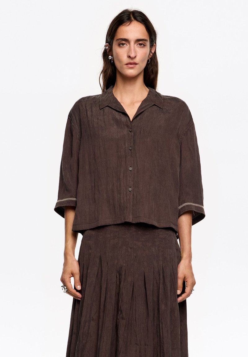 Brązowa, pomarszczona koszula z tkaniny z kołnierzykiem, krótkimi rękawami i zapięciem na guziki. Charakterystyczne przeszycie przy mankiecie rękawa.