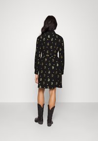 Musta mekko, jossa on kultainen paisley-kuvio, pitkät hihat, vyötärölle sidottu vyö ja polvipituinen helma, yhdistettynä mustiin cowboy-saappaisiin.