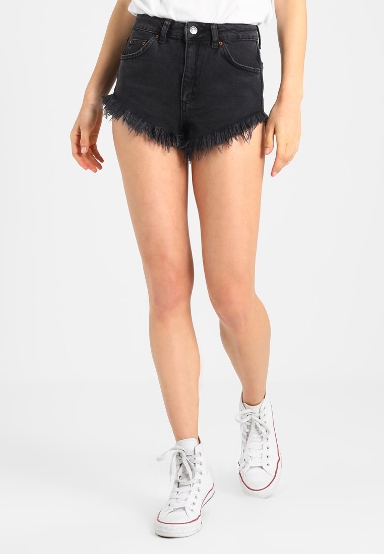Kvinna klädd i högmidjade svarta denimshorts med fransade kanter och vita högskaftade sneakers, stående mot en enkel vit bakgrund.