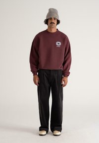 Burgundský sweatshir s vyšitým logom, kombinovaný s texturovanými čiernymi nohavicami a krémovými teniskami. Model má na sebe šedú pletenú čiapku.