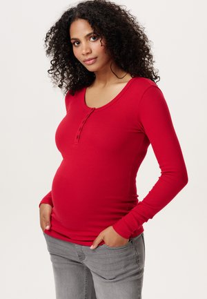 Schwangere Frau mit lockigem Haar, die ein rotes Langarmshirt und graue Jeans trägt, steht mit den Händen in den Taschen vor einem schlichten Hintergrund.