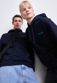 Des sweat-shirts bleu marine avec cordons de serrage et logo "tommyjeans" brodé en bleu-vert, associés à un jean en denim bleu clair. Texture lisse, coupe décontractée.