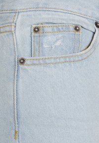 Os jeans em denim azul claro apresentam um pequeno bolso frontal com uma borda desfiada, hardware prateado e costura amarela ao longo das costuras.