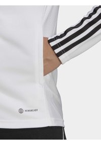 Hand in der Tasche eines weißen Langarmoberteils mit schwarzem Adidas-Drei-Streifen-Detail und "AEROREADY"-Logo nahe dem Saum.