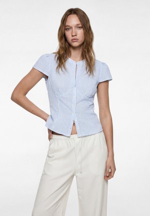 Young woman with long brown hair wearing a blue and white striped short-sleeve buttoned shirt and white pants against a plain background.