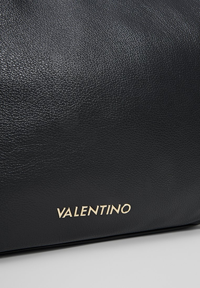 Surface en cuir noir texturé avec le logo doré en métal "VALENTINO" embossé près du bord inférieur sur un fond gris clair.