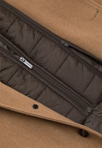 Brązowy wełniany płaszcz wewnątrz ma pikowaną, brązową podszewkę z zamkami, która charakteryzuje się gładką teksturą i funkcjonalnym designem. Posiada dwie kieszenie.