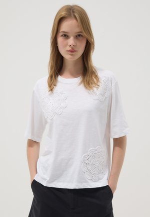 Young woman with shoulder-length blonde hair wearing a white T-shirt featuring three large white crochet patches, hands in black pants pockets.