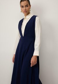 Rochie lungă bleumarin cu decolteu adânc în V, talie ajustată și fusta evazată. Purtați-o peste o cămașă crem cu nasturi. Cercei aurii ca accente notabile.