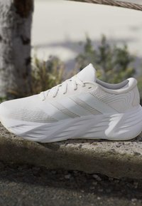Λευκό αθλητικό παπούτσι Adidas με τρεις ρίγες, παχιά μαξιλαρωτή σόλα και κορδόνια, τοποθετημένο πάνω σε πέτρινη επιφάνεια σε εξωτερικό χώρο.