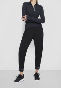 Žena oblečená v černé zipové dlouhé mikině a černých joggingových kalhotách s manžetami a brandingem Calvin Klein, stojící na bílém pozadí.