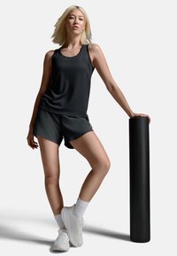 Sort atletisk tanktop og shorts, hvide sneakers, stående ved en tekstureret sort skumrulle med en neutral baggrund.