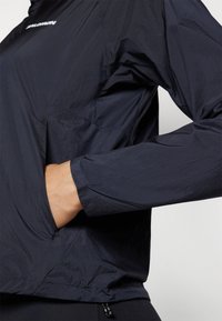 Lätt svart jacka med en blank yta, en ficka med dragkedja och elastiska ärmslut; har en halvdragkedja vid kragen och en avslappnad passform.