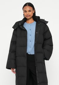 Lang sort pufferjakke med quiltet design, en aftagelig hætte og knapdetaljer, båret over en lys blå skjorte.