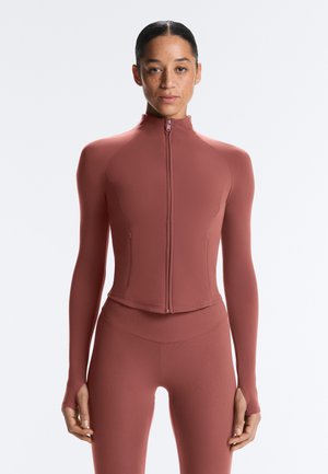 Nő hátrafésült hajjal, visel szűk szabású, hosszú ujjú cipzáras felsőt és hozzáillő leggingset tompa terrakotta színben, egyszerű háttér előtt állva.