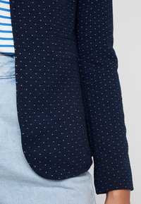 Granatowa marynarka o fakturowanej tkaninie, zdobiona białymi kropkami, o dopasowanym kroju i lekko zaokrąglonych mankietach.