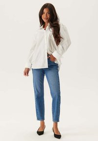 Vit skjorta med långa ärmar, parat med blå denim jeans med rak ben. Modell i svarta spetsiga klackskor. Neutral bakgrund.
