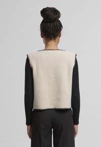 Béžový fleecový vest s černými detaily, obdélníkový tvar, otevřená záda, nošený přes černé dlouhé rukávy, vystavený na neutrálním pozadí.