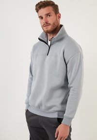 Sudadera de cremallera gris claro con cuello alto, hecha de una tela suave. Presenta puños y dobladillo de canalé, y un detalle de cremallera negra.