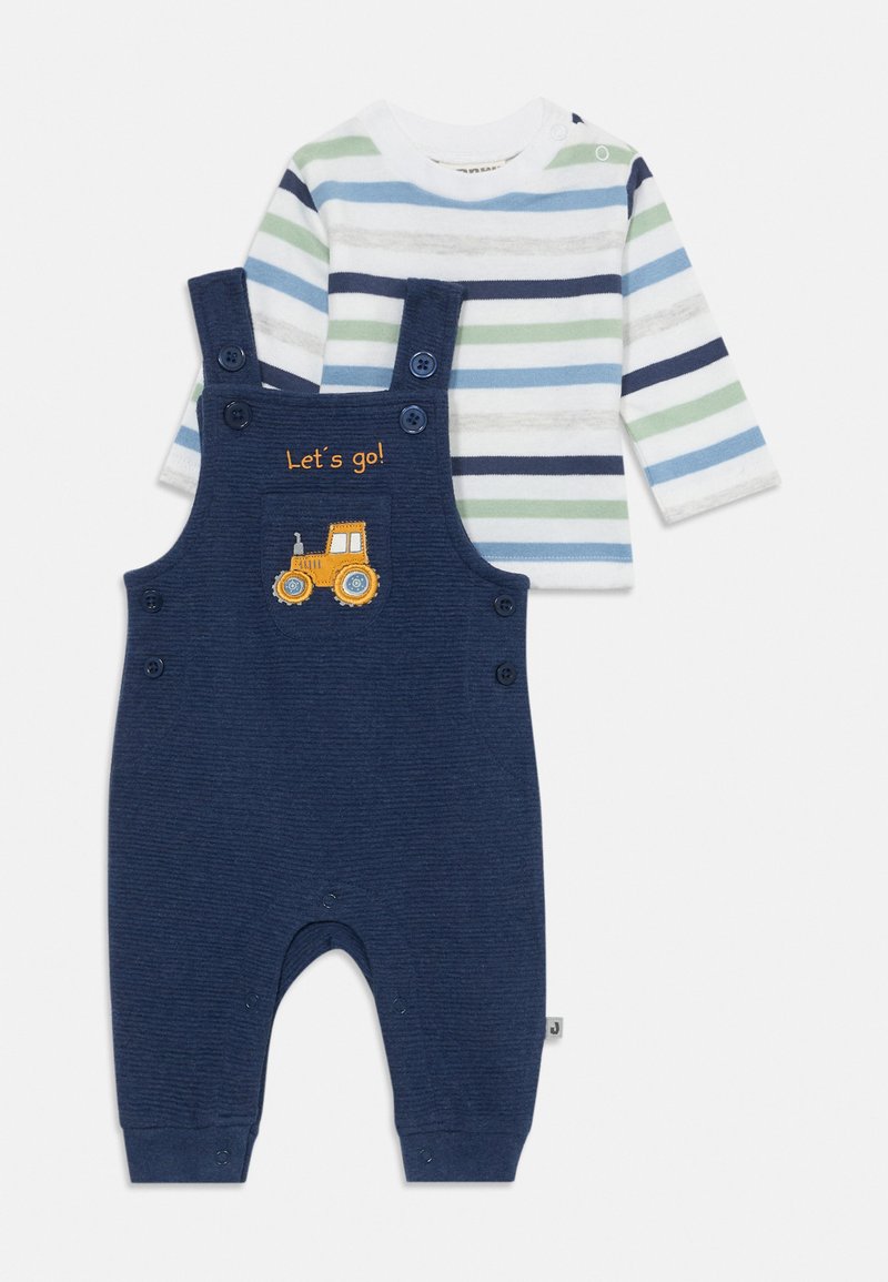 Salopette blu navy con texture a coste e design di un trattore giallo ricamato; abbinata a una maglietta a maniche lunghe a righe bianca, blu e verde.