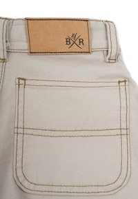 Beige denimshorts med en tan läderpatch vid midjan som har en logotyp. Bakfickan är synlig med bruna sömmar och en texturerad yta.