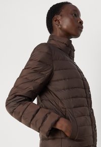 Veste marron matelassée avec un col haut, des coutures horizontales et des fermetures à bouton-pression. Le matériau semble lisse et léger.