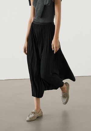 Kvinde iført grå ærmeløs top med matchende sweater draperet over skuldrene, sort plisseret midi nederdel og lysebeige snøresko.