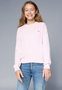 Hellrosa, Kabelstrickpullover mit gerippten Bündchen und Saum. Kleines Logo-Detail auf der Vorderseite. Über blauen Jeans getragen.