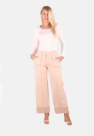 Frau mit langen blonden Haaren, die ein weißes langärmeliges Oberteil, beige weite Hosen und beige offene Sandaletten trägt, steht mit den Händen in den Taschen.