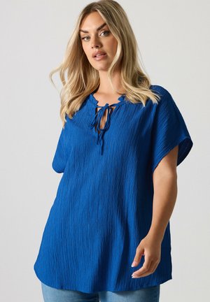 Blonde woman wearing a textured royal blue blouse with short sleeves and a front tie neckline, paired with light blue jeans.