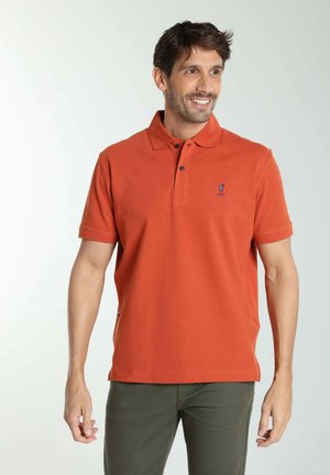 Homme portant un polo orange à manches courtes et un pantalon vert olive, souriant et regardant vers la droite sur un fond clair uni.