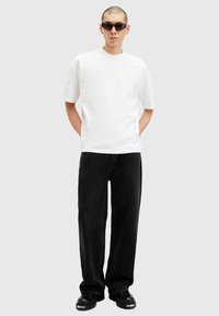 White cotton t-shirt featuring embossed text, paired with black loose-fit jeans and chunky black shoes. Accessorized with dark sunglasses.