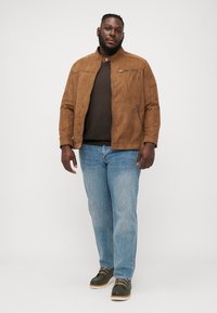 Veste en daim marron avec un col montant, poche zippée et deux poches latérales, portée sur une chemise foncée et un jean bleu clair.