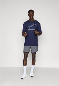 Navy blue Under Armour t-shirt with silver logo, paired with gray shorts and white athletic shoes. Individual holds a jump rope.