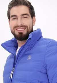 Blå puffy-jacka med dragkedja, hög krage och en diskret logotyp. Slät textur med ett kviltat mönster och kontrasterande dragkedjedetalj.