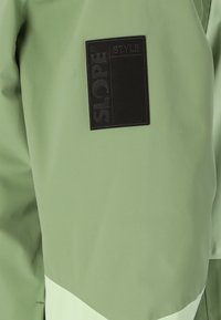 Vihreä vedenpitävä takki, jossa on tekstuurilla varustettu musta logo-ommel. Ominaisuuksiin kuuluu kerroksellinen muotoilu, vaaleampi alareuna ja sileä pinta.