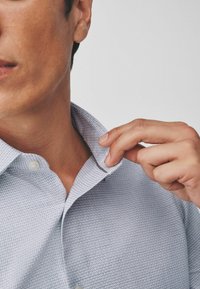 Light blue, textured button-up shirt with a crisp collar, showing a subtle pattern. A hand is adjusting the collar near the neck.
