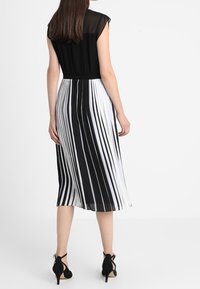 Schwarzes Oberteil mit transparenten Ärmeln, das in einen Midikleid übergeht, das abwechselnd schwarze und weiße senkrechte Streifen hat. Schwarze High Heels.