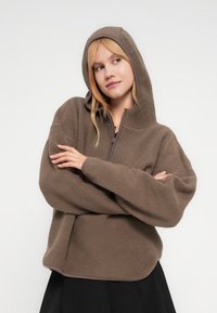 Καφέ φλις μπλούζα με κουκούλα, με φερμουάρ στη μέση και αντίθετες λεπτομέρειες. Το ύφασμα έχει απαλό, υφασμάτινο φινίρισμα.