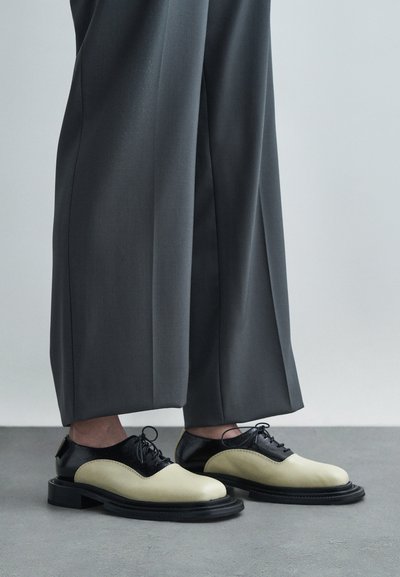 Chaussures Oxford bicolores noir et crème avec des bouts ronds et une épaisse semelle noire, accompagnées de pantalons gris amples.