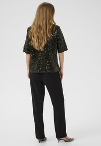 Melnas sekvīnēm rotāta blūze ar īsām piedurknēm, kombinēta ar melniem biksēm un svītrainiem augstpapēžu apaviem, skatīta no aizmugures.