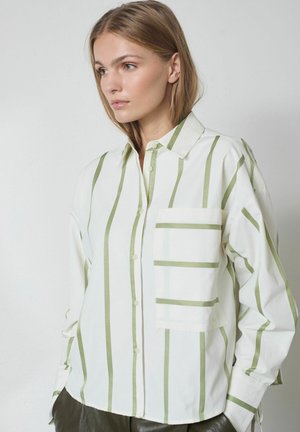Junge Frau trägt eine weiße Hemdbluse mit vertikalen grünen Streifen und einer großen Tasche auf der Brust, schaut leicht zur Seite.