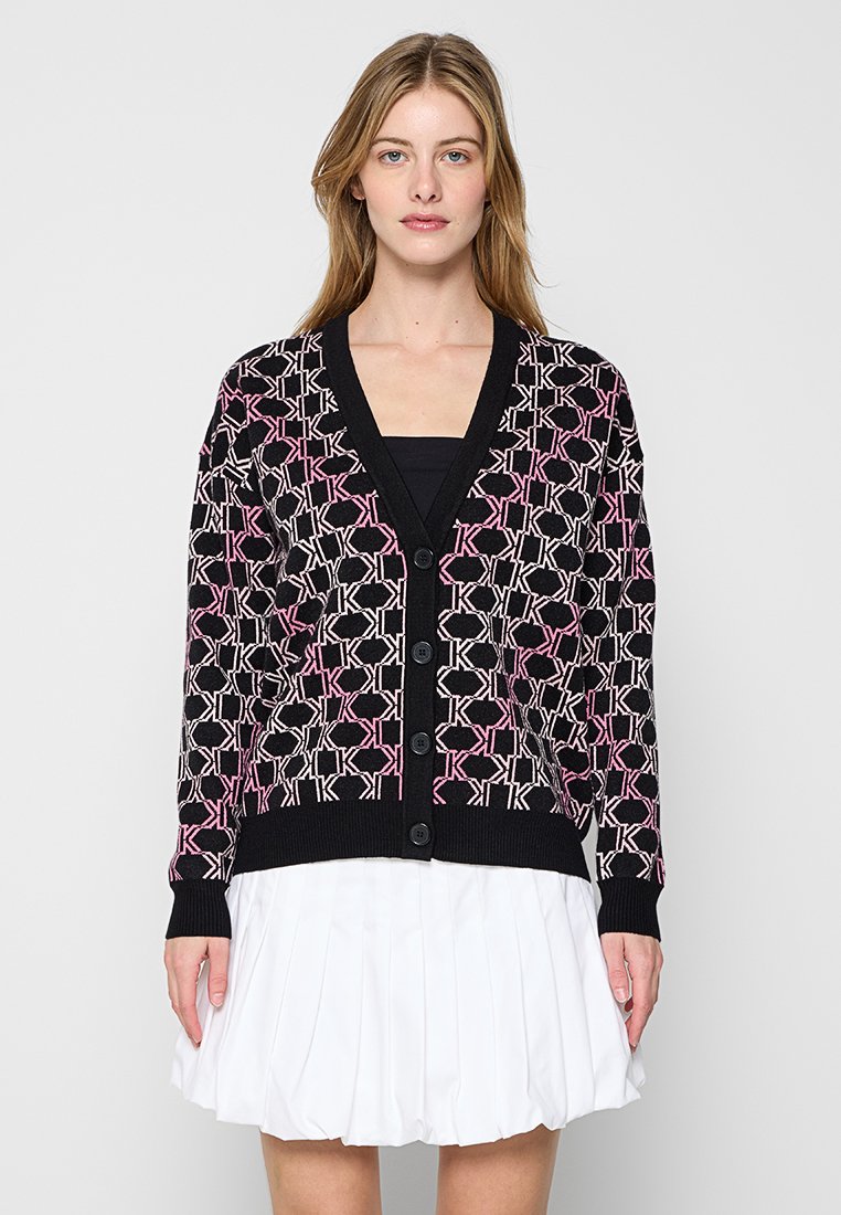 Schwarze Strickjacke mit einem geometrischen Muster in Pink und Weiß, mit Knopfleiste vorne und gerippten Bündchen und saum. Getragen über einem schwarzen Oberteil.