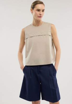 Frau trägt ärmelloses beiges Oberteil und weite marineblaue Shorts, die Hände in den Taschen, schaut leicht nach rechts vor einfarbigem Hintergrund.