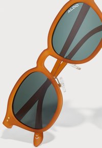 Gafas de sol mate naranjas con lentes cuadrados de gran tamaño, tinte verde oscuro y acentos metálicos. Presenta almohadillas nasales transparentes y un diseño de ceja plana.